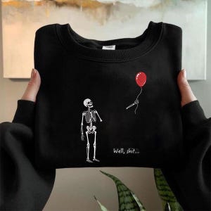 May include: A black crewneck sweatshirt featuring a graphic print of a white skeleton looking up at a red balloon held by a skeletal hand. The text "Well, shit..." is printed in white below the skeleton on the front of the garment.