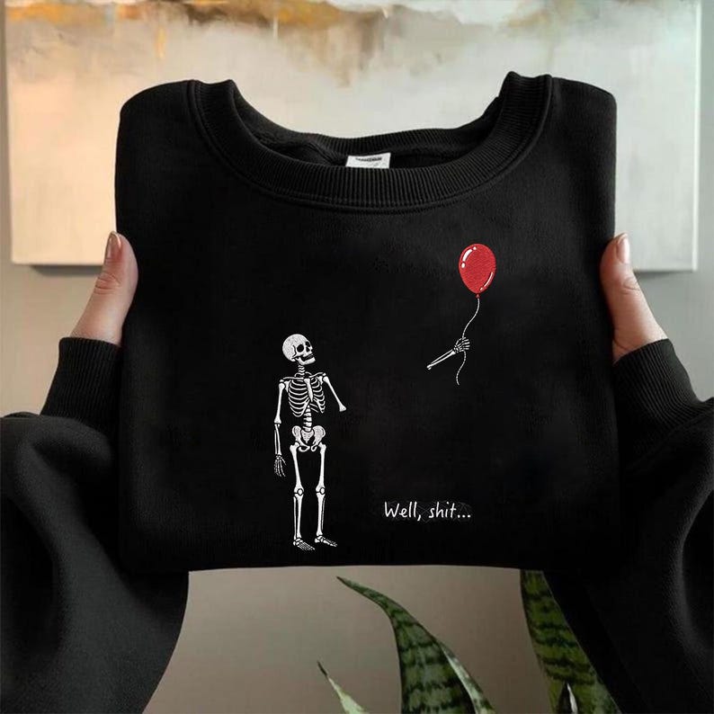 May include: A black crewneck sweatshirt featuring a graphic print of a white skeleton looking up at a red balloon held by a skeletal hand. The text "Well, shit..." is printed in white below the skeleton on the front of the garment.