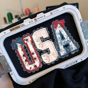 Op de afbeelding: Geborduurde letters "USA" op zwarte stof, gehouden in een borduurring. De letters hebben bloempatronen, kanten randen en strikken. De "U" en "S" hebben bloemmotieven met rode randen, terwijl de "A" een blauw bloemmotief heeft.