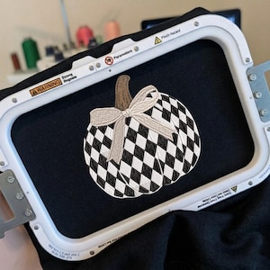 May include: An embroidery hoop frame holding black fabric with a stitched pumpkin design. The pumpkin features a black and white checkered pattern, a brown stem, and a light-colored ribbon bow tied around the stem, centered on the fabric.