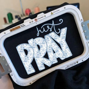 May include: A white rectangular embroidery hoop securing black fabric with the embroidered text "just PRAY". The word "just" is in a white cursive script, while "PRAY" features block letters filled with a blue and white floral pattern.