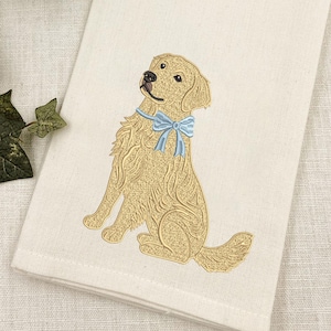 May include: A cream-colored kitchen towel featuring an embroidered golden retriever dog in a seated position. The dog is depicted in shades of gold and brown with a light blue ribbon tied around its neck. The towel has a textured fabric finish.