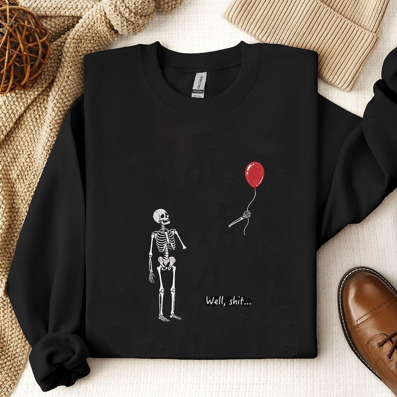 May include: A black long-sleeve sweatshirt featuring a graphic of a white skeleton standing and looking up at a red balloon held by a detached skeletal hand. The text "Well, shit..." is printed in white below the skeleton on the front of the garment.