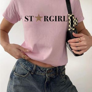 Puede incluir: Una camiseta de manga corta de color rosa claro con el texto negro STARGIRL impreso en el pecho, donde una estrella con estampado de leopardo sustituye a la letra A. La camiseta se combina con vaqueros azules y un bolso negro con clip.