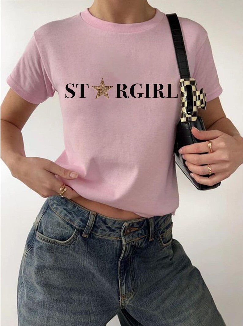 Puede incluir: Una camiseta de manga corta de color rosa claro con el texto negro STARGIRL impreso en el pecho, donde una estrella con estampado de leopardo sustituye a la letra A. La camiseta se combina con vaqueros azules y un bolso negro con clip.