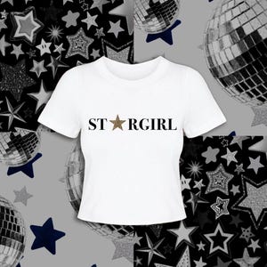 Puede incluir: Una camiseta blanca de manga corta con el texto negro "STARGIRL" impreso en el pecho, donde la letra "A" est&aacute; sustituida por una estrella de cinco puntas con un estampado de leopardo marr&oacute;n y negro en su superficie.