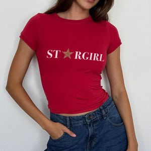 Puede incluir: Una camiseta roja de manga corta con el texto blanco STARGIRL impreso en el pecho, donde una estrella con estampado de leopardo marr&oacute;n reemplaza la letra A. La camiseta se combina con unos vaqueros azul oscuro con un bot&oacute;n y bolsillo visibles.