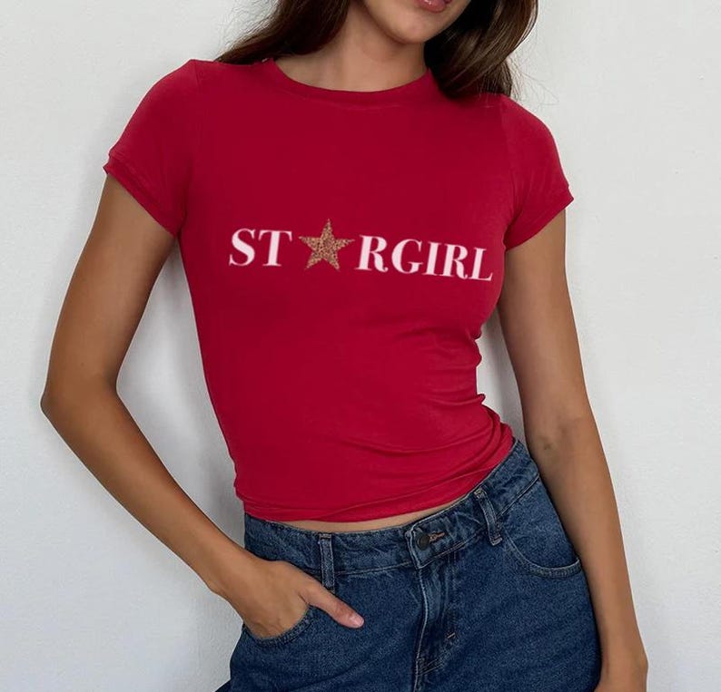 Puede incluir: Una camiseta roja de manga corta con el texto blanco STARGIRL impreso en el pecho, donde una estrella con estampado de leopardo marr&oacute;n reemplaza la letra A. La camiseta se combina con unos vaqueros azul oscuro con un bot&oacute;n y bolsillo visibles.