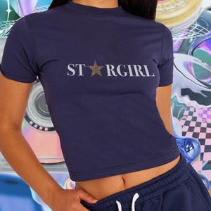 Puede incluir: Una camiseta de manga corta en color azul marino oscuro con el texto STARGIRL impreso en blanco en el pecho, donde la letra A es reemplazada por una estrella con estampado de leopardo en tono dorado. Tiene cuello redondo y corte corto.