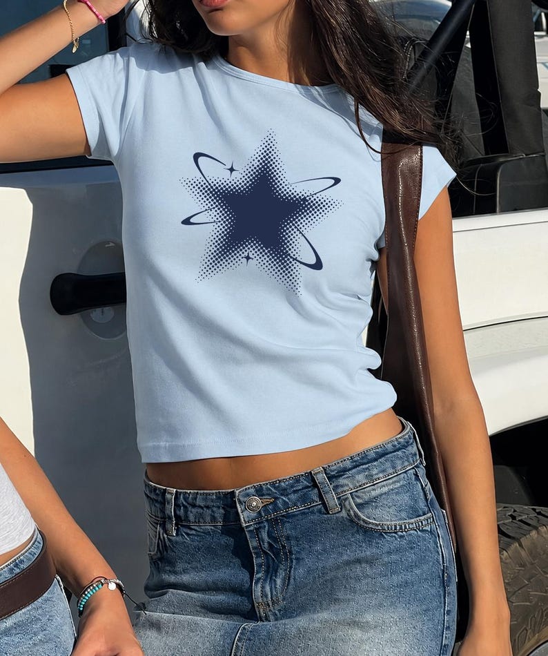 May include: A light blue short-sleeved t-shirt featuring a dark blue star graphic in the center. The star design is composed of a dotted halftone pattern and is surrounded by two thin, curved orbital lines with small four-pointed star accents.