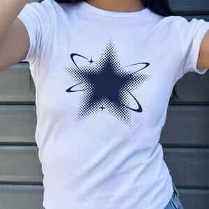 May include: A white short-sleeved t-shirt featuring a dark blue graphic of a five-pointed star. The star design incorporates a halftone dot pattern and is surrounded by two curved orbital rings and two small four-pointed star accents.