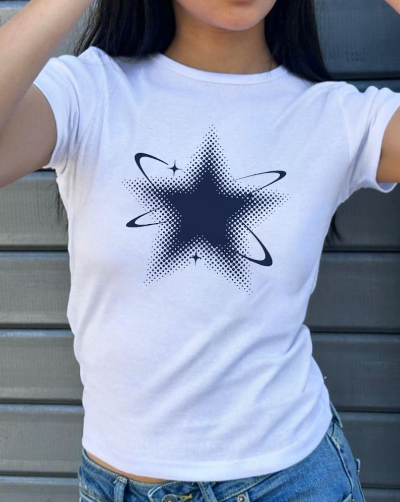 May include: A white short-sleeved t-shirt featuring a dark blue graphic of a five-pointed star. The star design incorporates a halftone dot pattern and is surrounded by two curved orbital rings and two small four-pointed star accents.