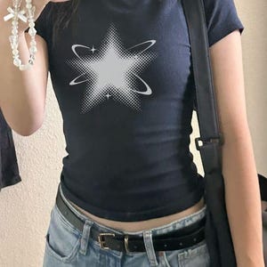 May include: A dark navy blue short-sleeved t-shirt featuring a white graphic of a star with a halftone dot pattern and orbiting rings. The shirt is worn with light blue denim jeans and a black belt, paired with a black shoulder bag.