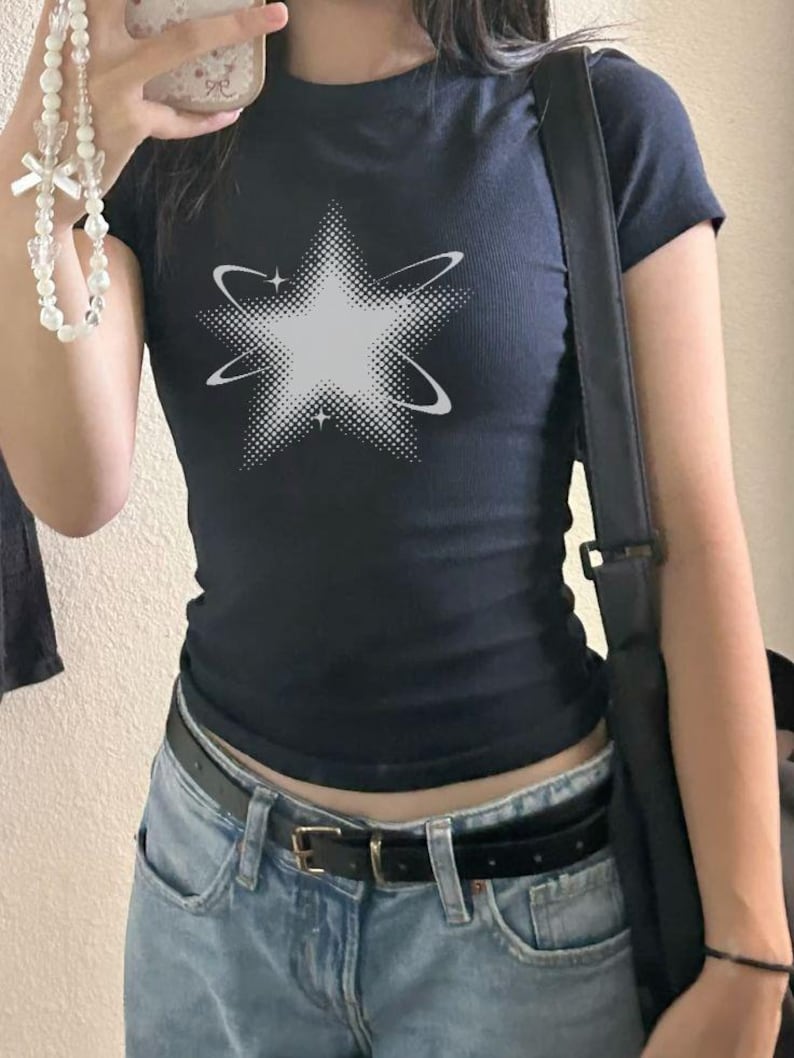 May include: A dark navy blue short-sleeved t-shirt featuring a white graphic of a star with a halftone dot pattern and orbiting rings. The shirt is worn with light blue denim jeans and a black belt, paired with a black shoulder bag.