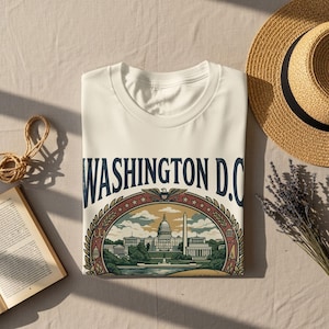 May include: Cream-colored t-shirt featuring a detailed illustration of Washington D.C. landmarks, including the Capitol Building and Washington Monument, within a decorative oval frame. The text "WASHINGTON D.C." is printed above the illustration in navy blue. A straw hat and a sprig of lavender are next to the shirt.