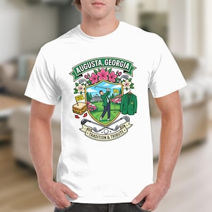 May include: White t-shirt featuring a colorful graphic with the text "AUGUSTA, GEORGIA". The design includes a golfer, flowers, a green jacket, and the words "TRADITION & TRIBUTE" on a banner. The shirt is worn by a person.