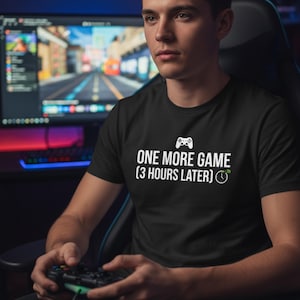May include: Black t-shirt with white text that reads "ONE MORE GAME (3 HOURS LATER)" and a controller icon. The shirt is worn by a person holding a game controller, with a computer monitor in the background.