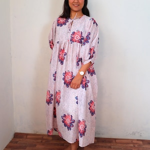 May include: A light pink dress with a floral pattern in red and blue. The dress has a loose fit, short sleeves, and a tie-neck detail. The floral design is repeated throughout the dress.