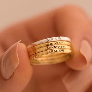 May include: A stack of four personalized rings. The top ring is silver with the word "LUCK" engraved. The other three rings are gold, with names "LINDSAY", "AMY&RYAN", and "FRANKIE" engraved. The rings are held in a person's hand.