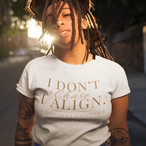 Puede incluir: Camiseta blanca con el texto dorado "I DON'T CHASE, I ALIGN.". La camiseta también presenta el texto "CONCIOUS COUTURE" y "520 7410". La persona lleva vaqueros azules.