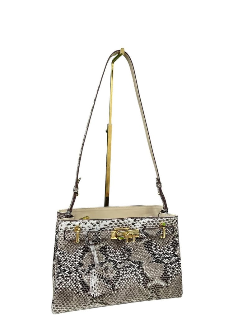 May include: A snakeskin-effect handbag with a gold-tone clasp and fittings. The bag features a top handle and a detachable shoulder strap. The interior is a light beige colour. The bag is displayed on a gold stand.