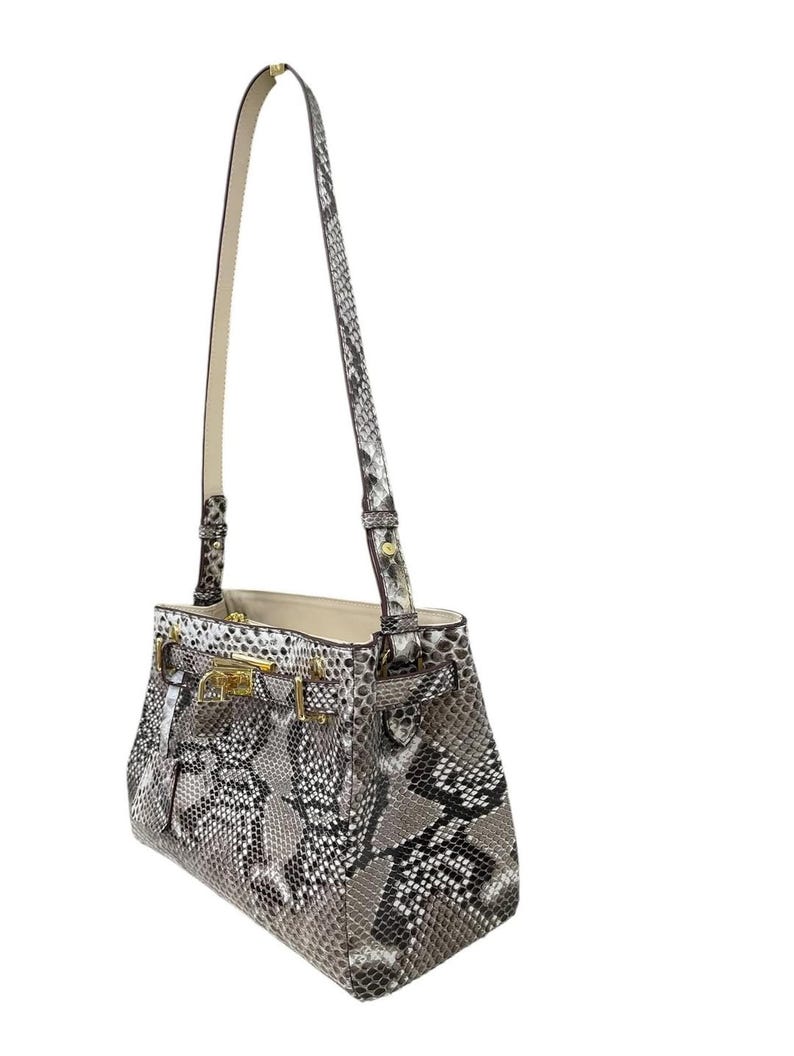 May include: A grey and white snakeskin-patterned handbag with a long, cream-coloured strap. The bag features gold-coloured hardware, including a clasp and buckles. The interior of the bag is a light beige colour.