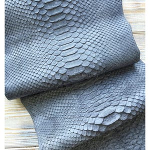 以下が含まれることがあります： グレーのヘビ革のクローズアップ。革は鱗模様で、折りたたまれています。鱗は中央が明るいグレーで、端が濃いグレーです。革は明るい色の木製の表面に置かれています。
