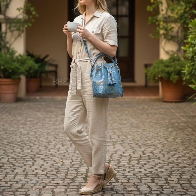 May include: A blue leather bucket bag with a textured finish and a top handle. The bag features a drawstring closure and a tassel. The bag is carried by a person wearing a beige jumpsuit.