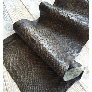 以下が含まれることがあります： 濃い灰色のヘビ革のクローズアップ。革は質感のある鱗模様で、折りたたまれており、明るい色の裏側が見えています。革は明るい色の木製の表面に表示されています。