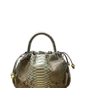 Könnte beinhalten: Eine silberfarbene Handtasche mit Kordelzug und oberem Griff. Die Tasche hat ein Schlangenmuster und goldfarbene Akzente an den Enden des Kordelzugs. Der Griff hat eine passende silberne Farbe.