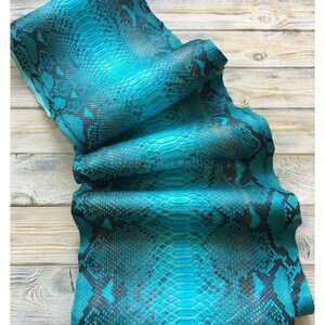 May include: A sheet of turquoise snakeskin leather with a scale pattern and dark grey markings. The leather is folded and draped over a light brown wooden surface. The text "exoticLeather" is visible.