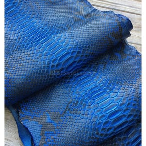 以下が含まれることがあります： 青色のエキゾチックレザーのクローズアップ。ヘビ革模様。 レザーは、青と黒の色合いのグラデーションを持つテクスチャ加工された表面を持っています。 素材は折りたたまれており、裏側が明らかになり、画像に奥行きが加わっています。 テキスト「BBSExoticLeather」が表示されています。