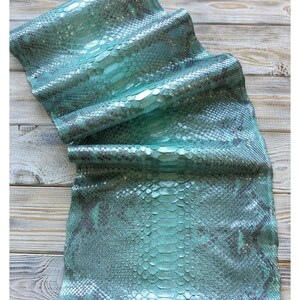 May include: A piece of teal-colored snakeskin leather with a scale pattern. The leather is folded and draped, revealing the texture and color variations. The background is a light-colored wooden surface.