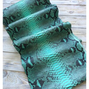 以下が含まれることがあります： ターコイズと黒色のヘビ革の折り畳まれた部分。革は鱗模様で、鮮やかなターコイズ色に黒い模様があります。素材は折りたたまれており、質感と模様を示しています。