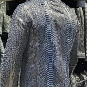 May include: A dark blue leather jacket with a snakeskin pattern. The jacket has a stand-up collar, long sleeves, and a zipper closure. The back of the jacket is visible, showcasing the intricate scale-like texture of the leather.