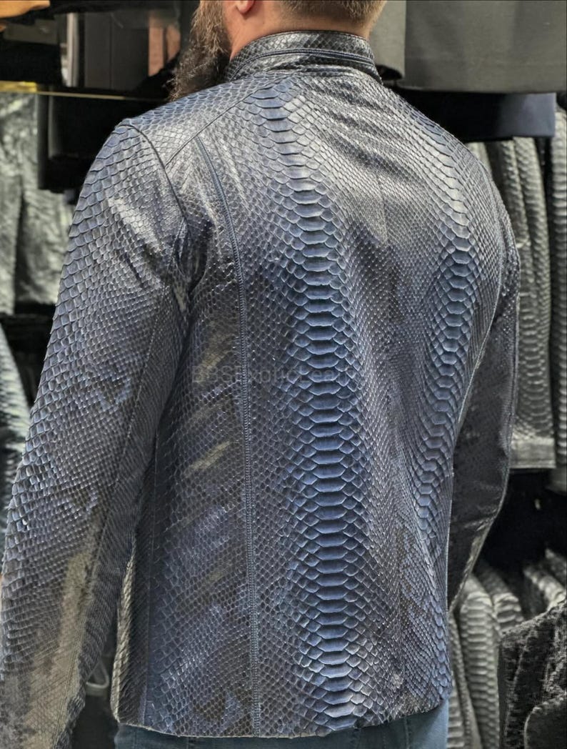 May include: A dark blue leather jacket with a snakeskin pattern. The jacket has a stand-up collar, long sleeves, and a zipper closure. The back of the jacket is visible, showcasing the intricate scale-like texture of the leather.