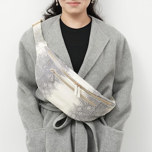 May include: A white and gray patterned fanny pack with two zippered compartments and gold-colored zippers. The bag has a curved shape and a wide strap. The fanny pack is worn across the body.