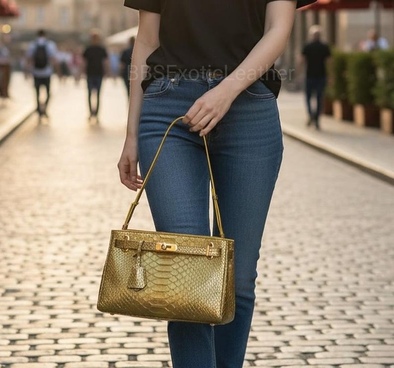 Puede incluir: Un bolso dorado con un estampado de piel de serpiente. El bolso tiene forma rectangular con un asa superior y un cierre de hebilla. El bolso lo lleva una persona con vaqueros azules y una camiseta negra. El texto "BBSE Exotic Leather" es visible.