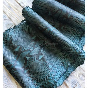 May include: Rolled pieces of teal and black snakeskin leather. The leather has a scale pattern and is arranged on a light wood surface. The text "exotic leather" is visible in the bottom left corner.