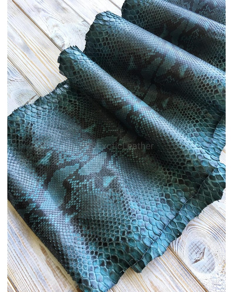 Peut inclure: Plusieurs rouleaux de cuir de peau de serpent bleu sarcelle et noir. Le cuir pr&eacute;sente un motif d'&eacute;cailles et est dispos&eacute; sur une surface en bois clair. Le texte "exotic leather" est visible en bas &agrave; gauche.