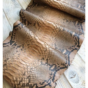 Può includere: Primo piano di una pelle di serpente conciata con un motivo dettagliato di squame marroni e nere. La pelle è piegata, rivelando la consistenza e i segni naturali. La pelle di serpente viene probabilmente utilizzata per l'artigianato.