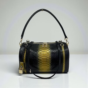 Könnte beinhalten: Schwarze und goldfarbene Handtasche mit Schlangenmuster, mit einem oberen Griff und einem abnehmbaren Schulterriemen. Die Tasche verfügt über goldfarbene Hardware, einschließlich Reißverschlüssen und einem kleinen Vorhängeschloss. Das Design umfasst ein zentrales Goldpaneel und schwarze Seitenteile.