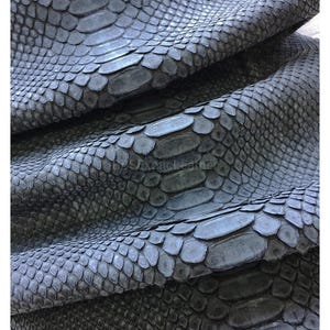 以下が含まれることがあります： グレーのヘビ革のシートのクローズアップ。革は、さまざまなグレーの色合いを持つ、質感のあるウロコ模様です。ウロコは重なり合うように配置され、自然で有機的な外観を作り出しています。画像は、折りたたまれ、ドレープされた革を示しています。