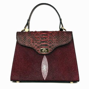 May include: A burgundy handbag with a textured finish and a contrasting snakeskin flap. The bag features a white stingray design on the front, a top handle, and gold-toned hardware. The bag is approximately 10 inches wide.