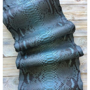 May include: A piece of teal-colored snakeskin leather with a scale pattern. The leather has dark blue and black accents, and the text "exotic leather" is visible. The leather is folded and draped over a light-colored wooden surface.