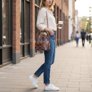 May include: A person walking on a city pavement, wearing a cream blouse, blue jeans, and white trainers. They carry a brown leather bucket bag with a snakeskin pattern in brown, purple, and pink. The bag has a drawstring closure and a tassel.