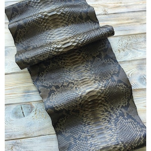 Op de afbeelding: Een opgerold stuk slangenleer met een schubbenpatroon in grijstinten, bruin en zwart. Het leer is gevouwen en gedrapeerd, wat de textuur en natuurlijke markeringen laat zien. De achtergrond is een lichtgekleurd houten oppervlak.