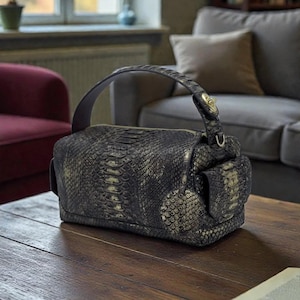 May include: A dark gray and gold snakeskin-patterned handbag with a top handle and a side pocket. The bag has a zipper closure and a gold-colored clasp on the handle. The bag is sitting on a wooden table.