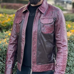 May include: A burgundy and brown leather jacket with a snakeskin pattern. The jacket has two chest pockets, a zipper closure, and a collared neckline. The sleeves and shoulders feature the snakeskin pattern, while the body is a solid brown color.