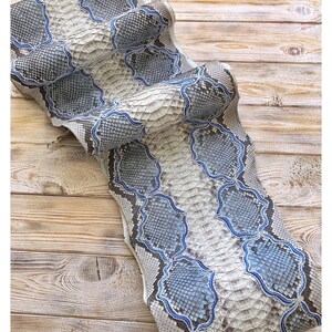 以下が含まれることがあります： グレー、白、青の色合いのヘビ革テキスタイル。ヘビ革の模様は、青で縁取られた大きな不規則な形が特徴です。このテキスタイルは、明るい色の木製の表面に表示されています。